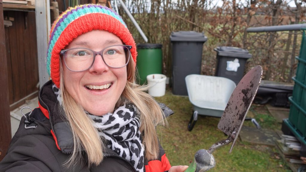 Maren lächelt vor dem Geräteschuppen im Garten, mit einer Schaufel in der Hand, bereit für die Gartensaison. Es geht um die Aussaat im Februar